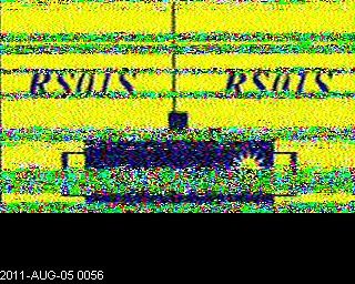 Slow-scan television image dominated by a yellow field on which are shown the satellite's shape and its callsign, RS01S, in dark blue. Many multicolored bands interrupt the image horizontally, reflecting noise due to varying signal strength.