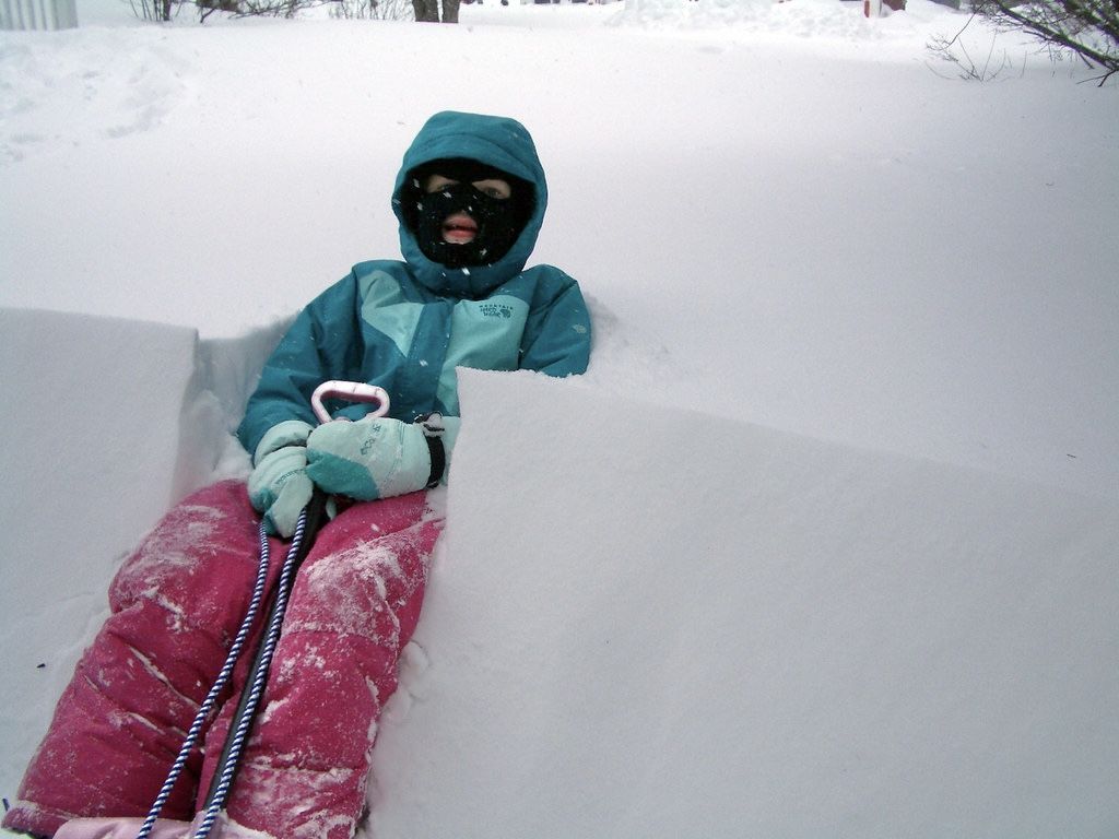 A small person in a pink and teal snow suit and wearing a face mask sits deep in a steep, peaked snowbank.