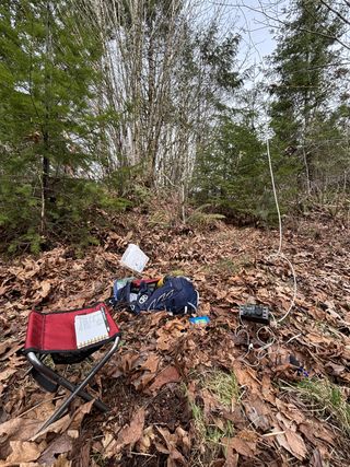 A red folding stool, blue backpack, and radio gear set up on a carpet of dried, brown leaves. Small trees are in the background. A white cable rises to a barely perceptible wire antenna.
